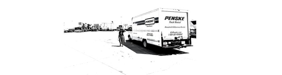 A black and white photo is shown of a woman wearing a hat, looking at the camera from a distance, next to a Penske moving truck, in a wide open parking lot.