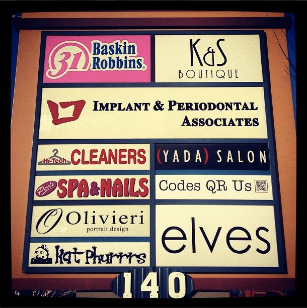 A strip mall marquee with the buiness logos is centered. From top, left to right, the following business names are shown: Baskin Robbins, K&S Boutique, Implant and Periodontal Associates, Hi-Tech Cleaners, Yada Salon, Pedi Spa & Nails, Codes QR Us, Olivieri Portrait Design, elves, and Kat Phurrrs.
